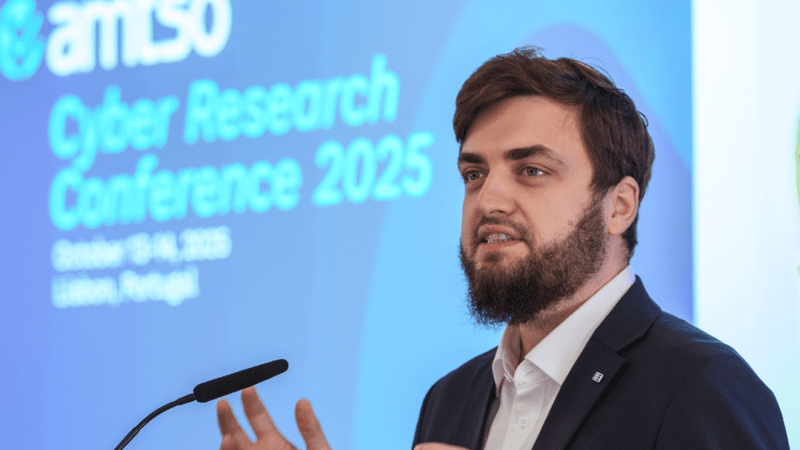 Vlad Iliushin, ELLIO Founder and AMTSO President presenting at cybersecurity conference with "Cyber Research Conference 2025" displayed on blue projection screen behind podium