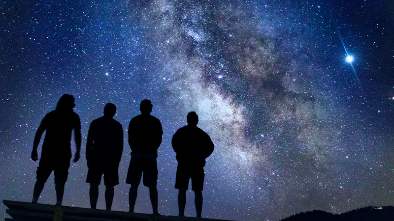 The image shows a group of people gazing at the night sky, where the Milky Way is visible.